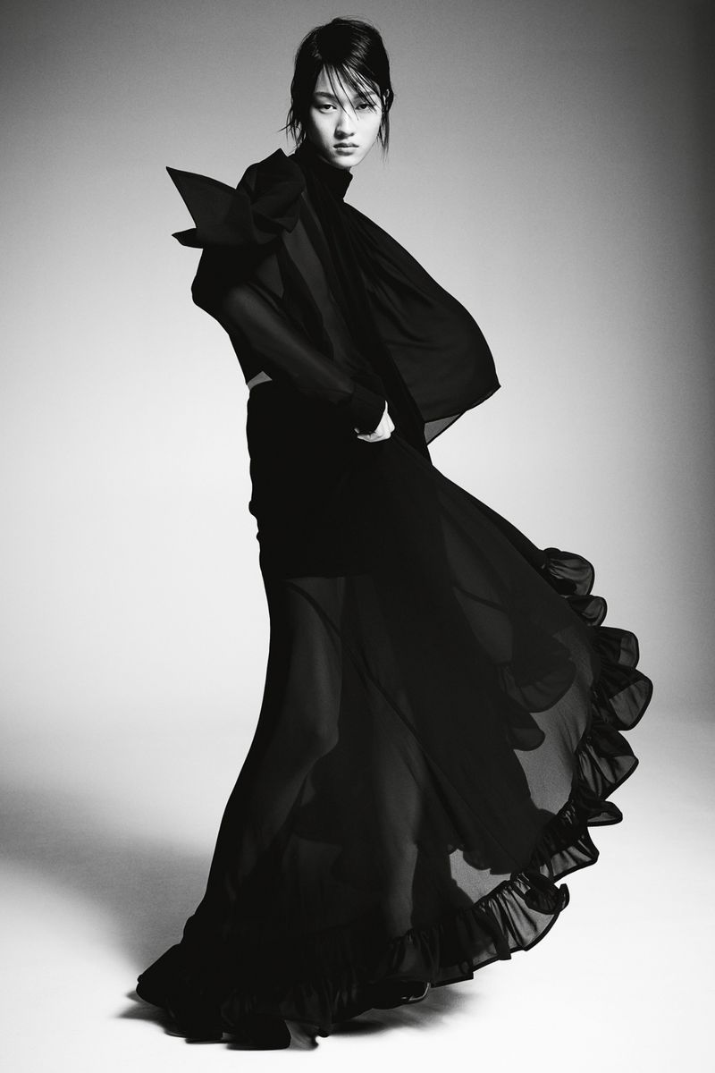 A fashionable woman poses in a dark, flowing dress with dramatic shoulder detail against a gradient studio backdrop, showcasing a stylish design.