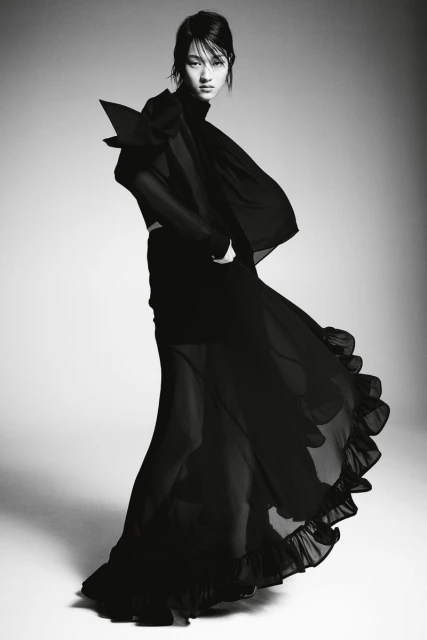 A fashionable woman poses in a dark, flowing dress with dramatic shoulder detail against a gradient studio backdrop, showcasing a stylish design.