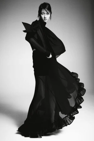 A fashionable woman poses in a dark, flowing dress with dramatic shoulder detail against a gradient studio backdrop, showcasing a stylish design.