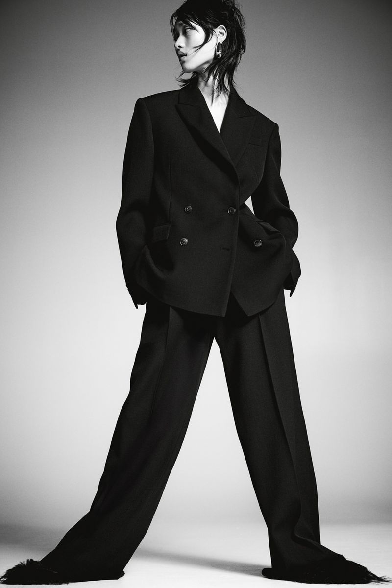 A striking fashion shot of a slender person wearing a dark double-breasted suit, standing against a neutral gradient background.
