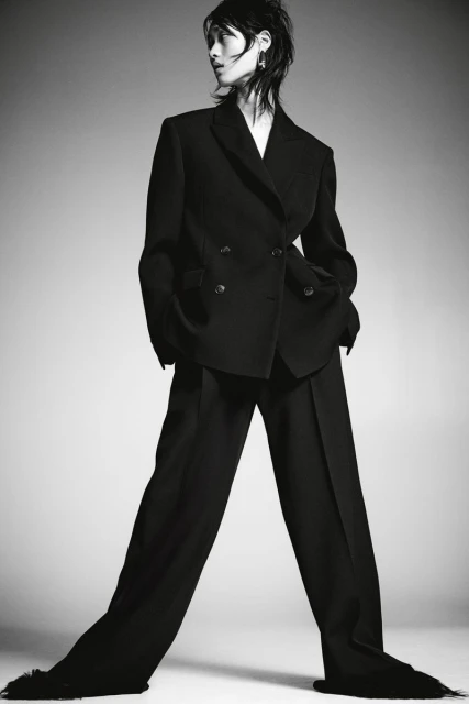 A striking fashion shot of a slender person wearing a dark double-breasted suit, standing against a neutral gradient background.
