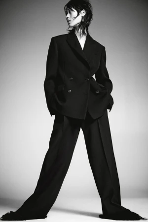 A striking fashion shot of a slender person wearing a dark double-breasted suit, standing against a neutral gradient background.