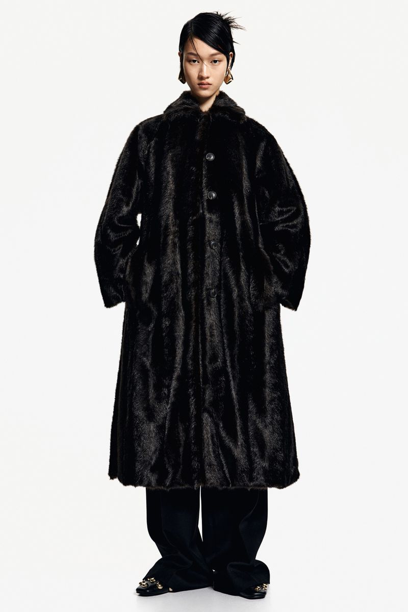 Asian fashion model wears a long black fur coat with black pants against a white backdrop in a studio fashion shot.