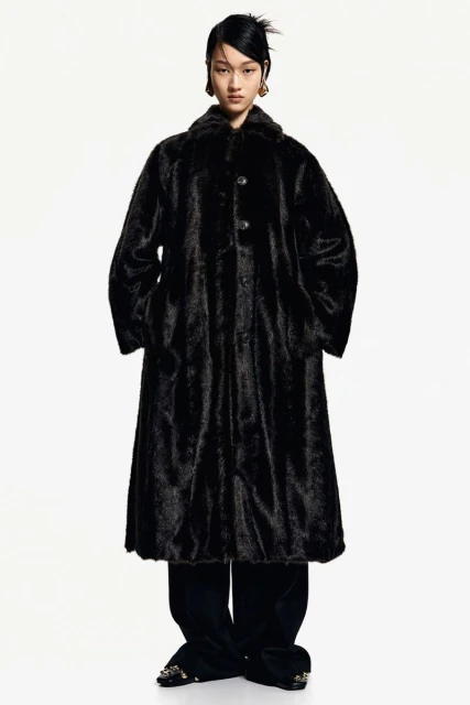 Asian fashion model wears a long black fur coat with black pants against a white backdrop in a studio fashion shot.