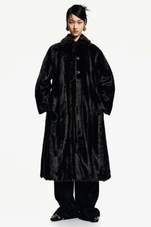 Asian fashion model wears a long black fur coat with black pants against a white backdrop in a studio fashion shot.