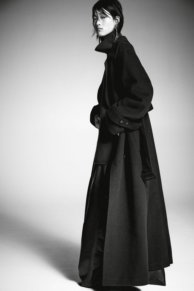 Model wearing a long black coat and high-neck sweater against a seamless light grey studio backdrop.