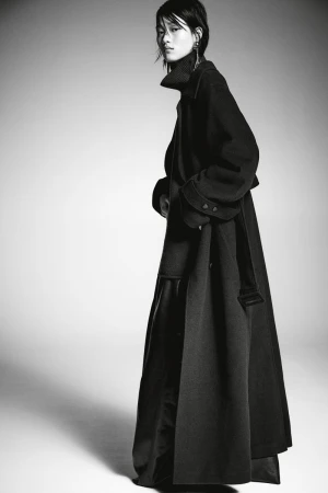 Model wearing a long black coat and high-neck sweater against a seamless light grey studio backdrop.