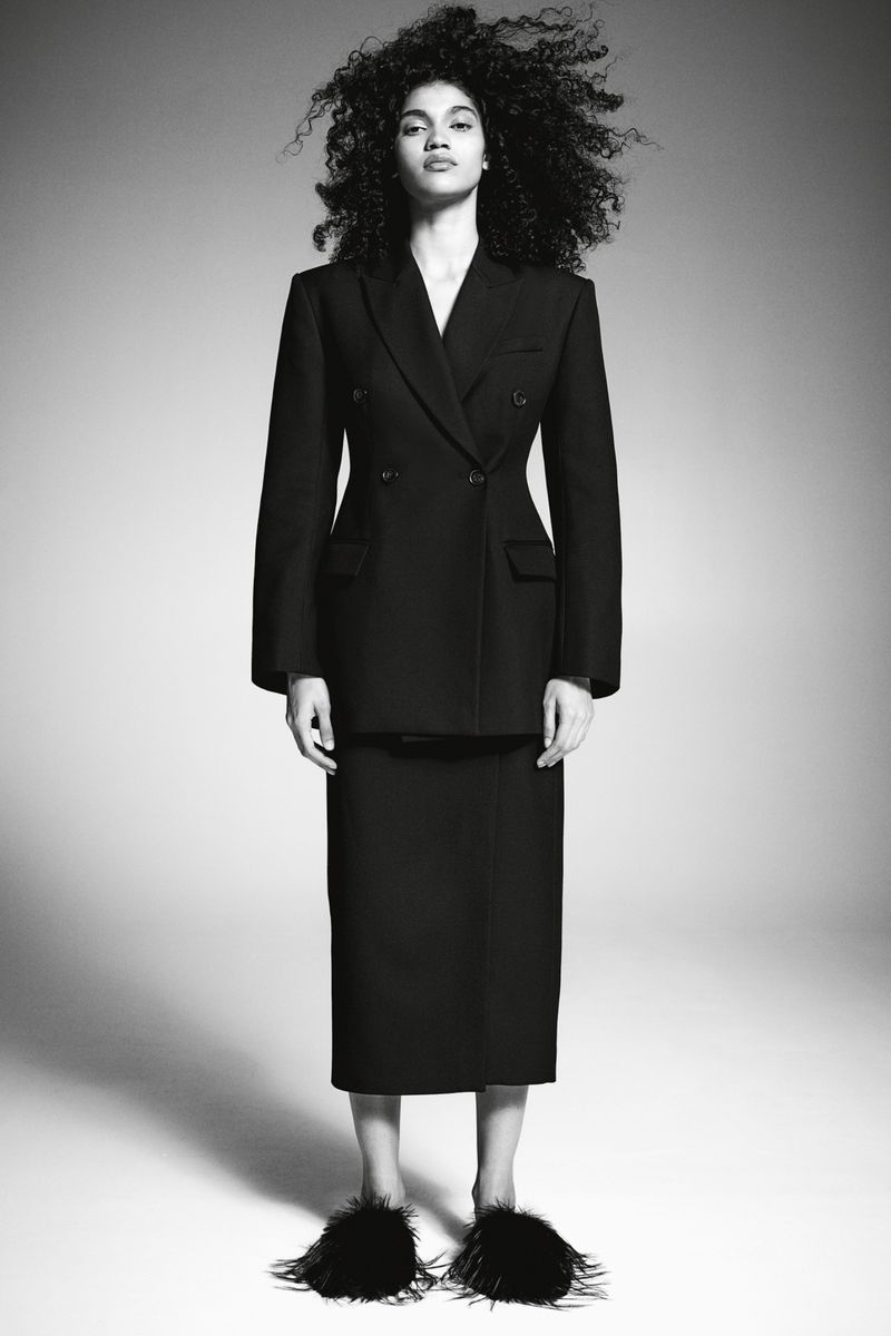 Full-length portrait of a woman with dark, curly hair wearing a dark skirt suit and black feather slippers on a neutral background.
