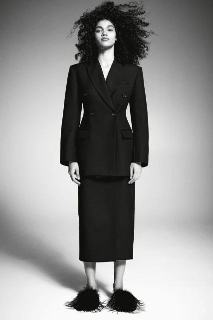 Full-length portrait of a woman with dark, curly hair wearing a dark skirt suit and black feather slippers on a neutral background.