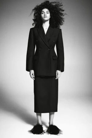 Full-length portrait of a woman with dark, curly hair wearing a dark skirt suit and black feather slippers on a neutral background.
