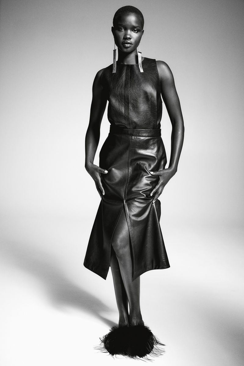 Black and white fashion portrait of a Black woman wearing a leather dress, statement earrings, and feathered slippers in a studio.
