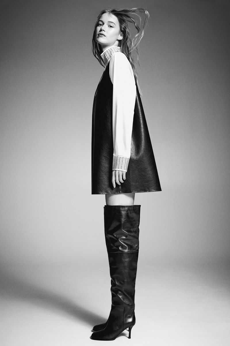 Black and white fashion shot of a woman in a black dress over a white shirt with tall black boots on a grey gradient background.