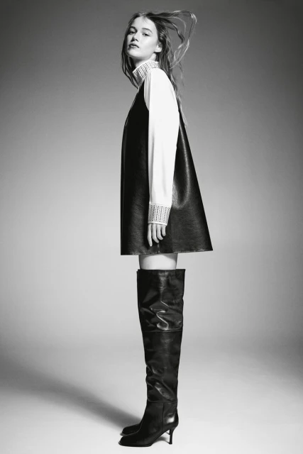 Black and white fashion shot of a woman in a black dress over a white shirt with tall black boots on a grey gradient background.