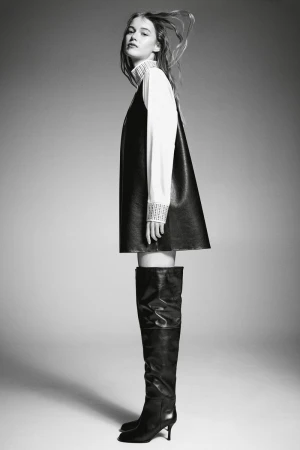 Black and white fashion shot of a woman in a black dress over a white shirt with tall black boots on a grey gradient background.
