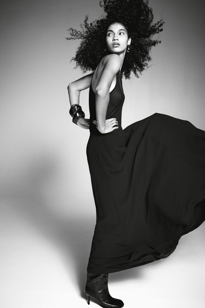Black and white fashion portrait of a woman in a gown and boots standing in a studio looking left with her hands on her hips.