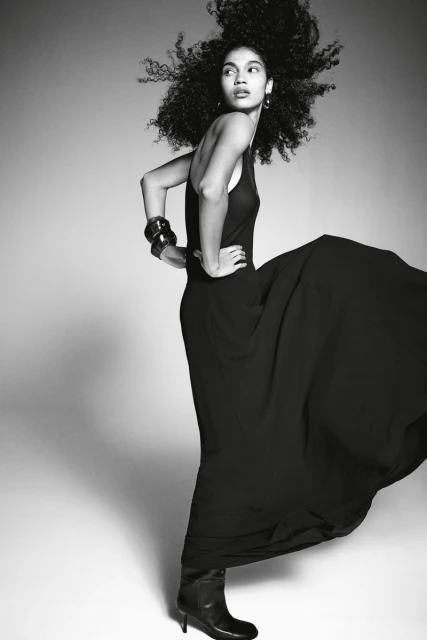 Black and white fashion portrait of a woman in a gown and boots standing in a studio looking left with her hands on her hips.