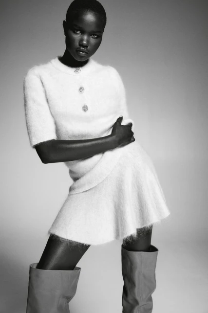Elegant African woman in a fuzzy white skirt-and-top set poses in gray thigh-high boots against a neutral background for a fashion editorial.