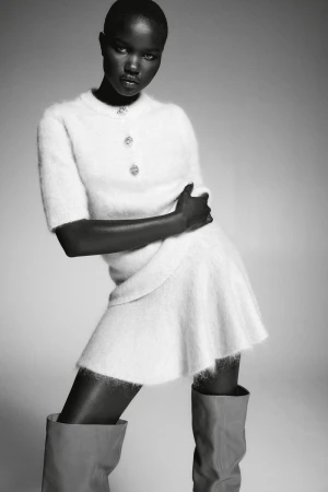 Elegant African woman in a fuzzy white skirt-and-top set poses in gray thigh-high boots against a neutral background for a fashion editorial.