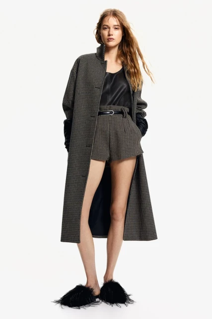 Fashion shot: Model poses in houndstooth coat and shorts, black top, and black slippers against a clean white backdrop in a studio setting.