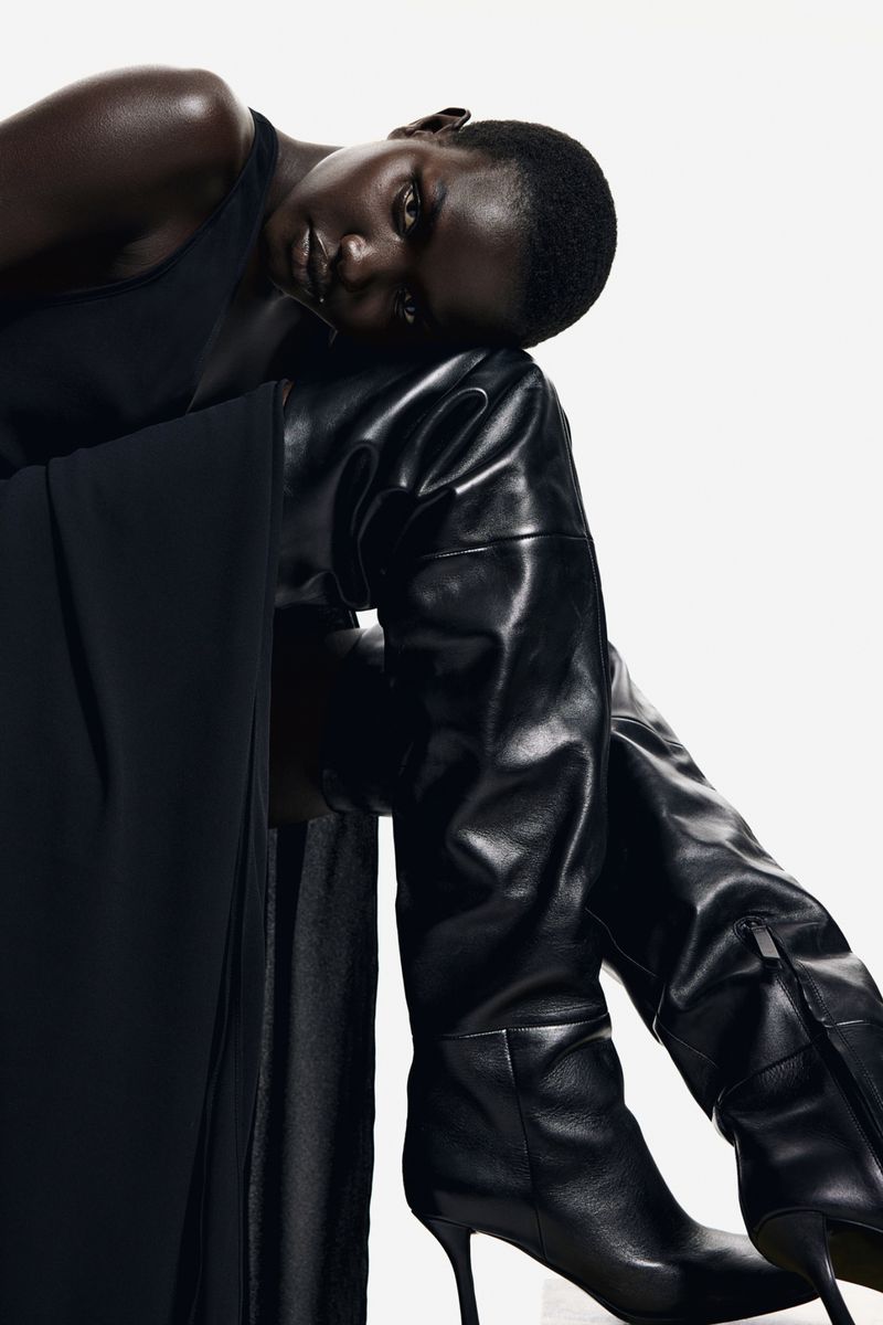 Elegant black woman with smooth skin poses in a dark dress and shiny, black, leather, high-heeled, thigh-high fashion boots against a white backdrop.