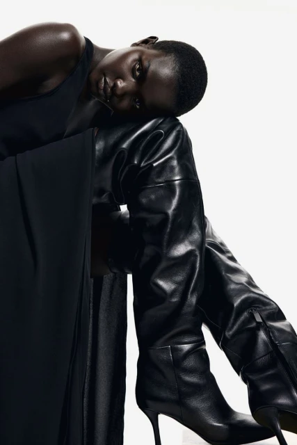 Elegant black woman with smooth skin poses in a dark dress and shiny, black, leather, high-heeled, thigh-high fashion boots against a white backdrop.