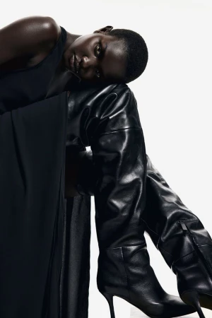 Elegant black woman with smooth skin poses in a dark dress and shiny, black, leather, high-heeled, thigh-high fashion boots against a white backdrop.