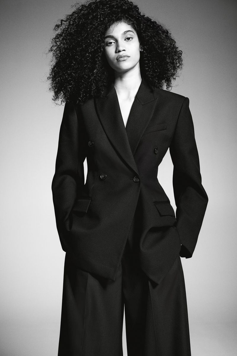 Fashion portrait of a young adult woman with curly hair, wearing a black double-breasted blazer and wide-leg pants against a grey backdrop.