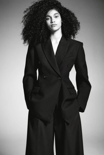 Fashion portrait of a young adult woman with curly hair, wearing a black double-breasted blazer and wide-leg pants against a grey backdrop.