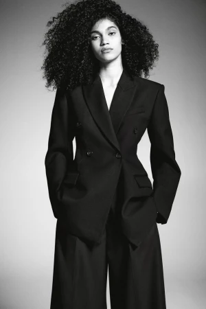 Fashion portrait of a young adult woman with curly hair, wearing a black double-breasted blazer and wide-leg pants against a grey backdrop.