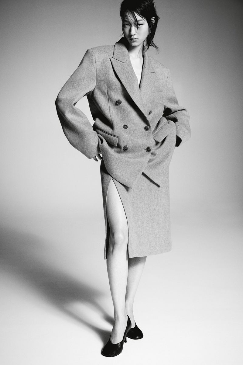 Asian woman posing in an oversized double-breasted grey wool suit and black leather heels against a neutral grey backdrop in studio lighting.