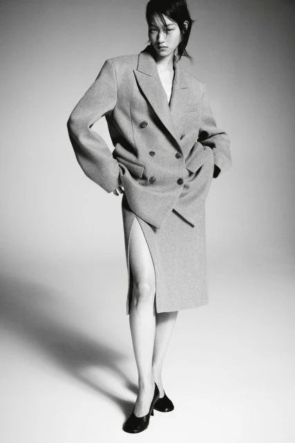 Asian woman posing in an oversized double-breasted grey wool suit and black leather heels against a neutral grey backdrop in studio lighting.