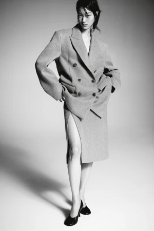 Asian woman posing in an oversized double-breasted grey wool suit and black leather heels against a neutral grey backdrop in studio lighting.