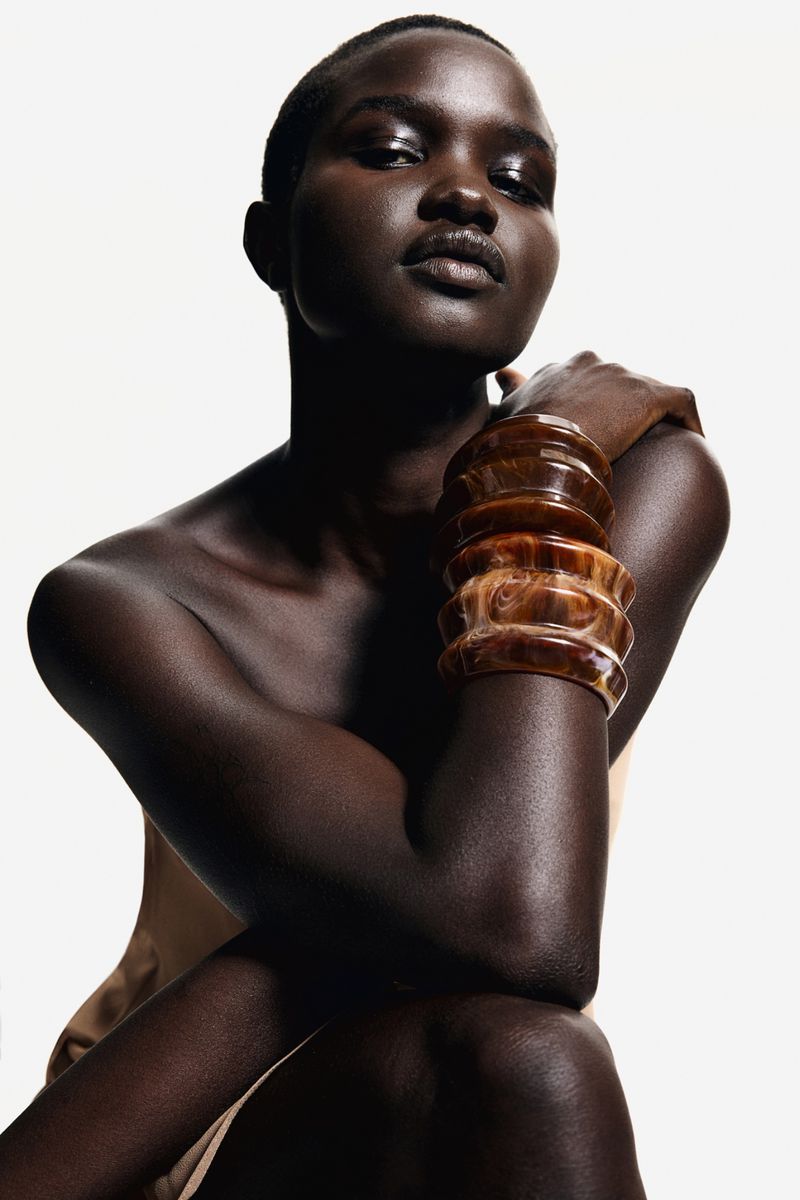 Woman with dark skin posing in a studio. She is wearing large amber-colored bangles on her arm and has a neutral facial expression.
