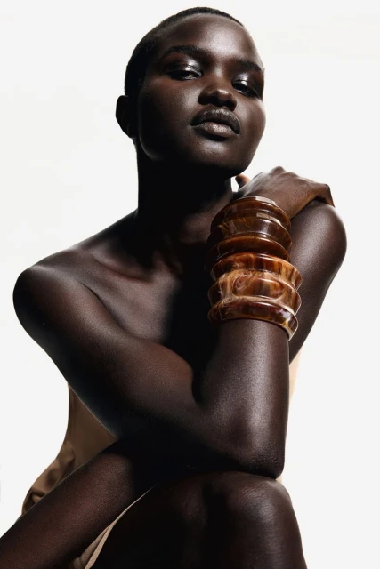 Woman with dark skin posing in a studio. She is wearing large amber-colored bangles on her arm and has a neutral facial expression.