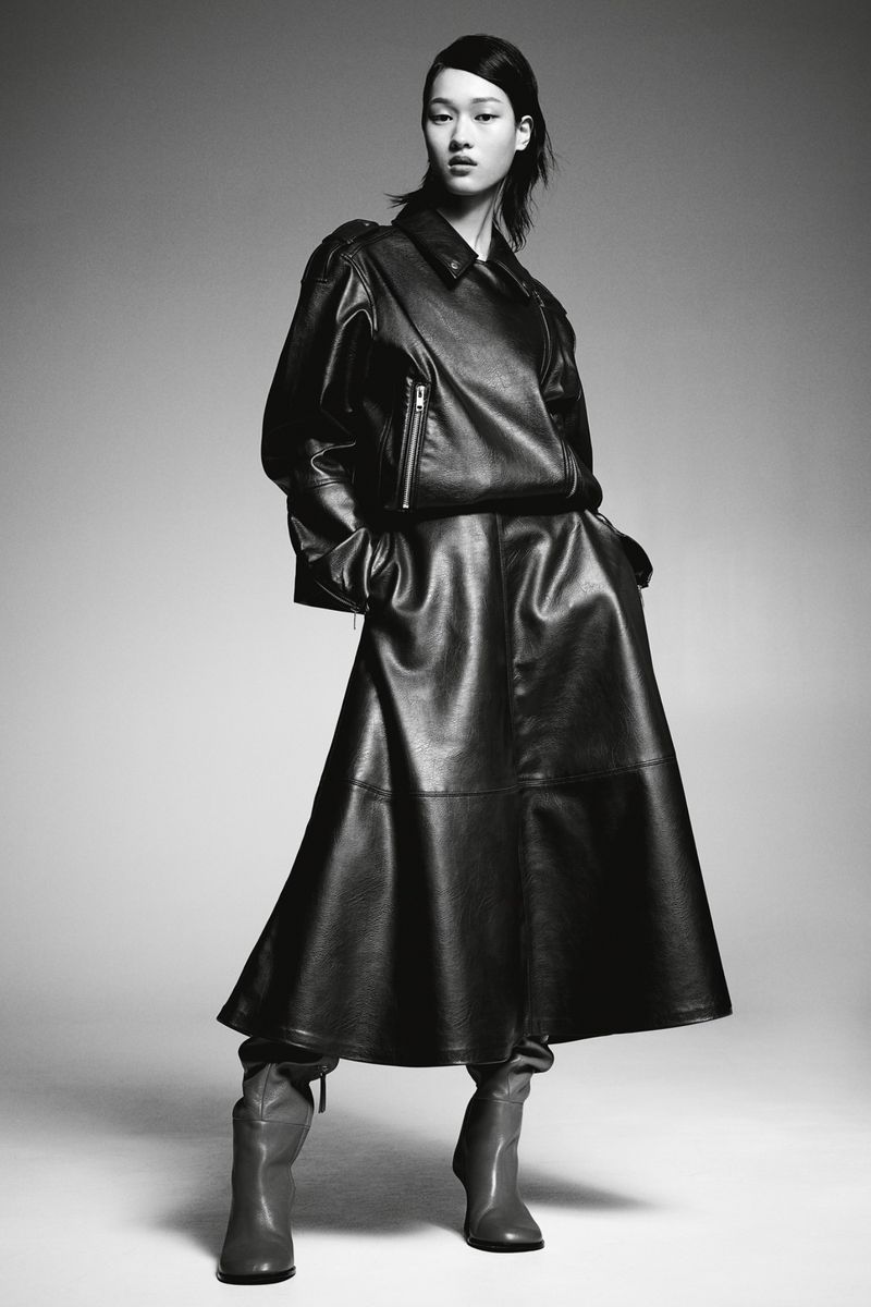 Elegant woman in a black leather dress, jacket, and boots strikes a confident pose against a studio backdrop.
