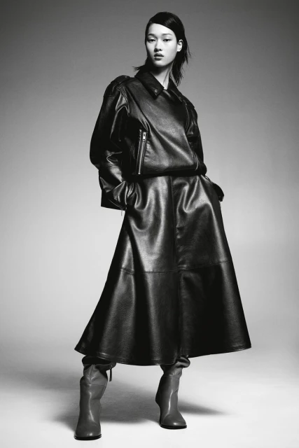 Elegant woman in a black leather dress, jacket, and boots strikes a confident pose against a studio backdrop.