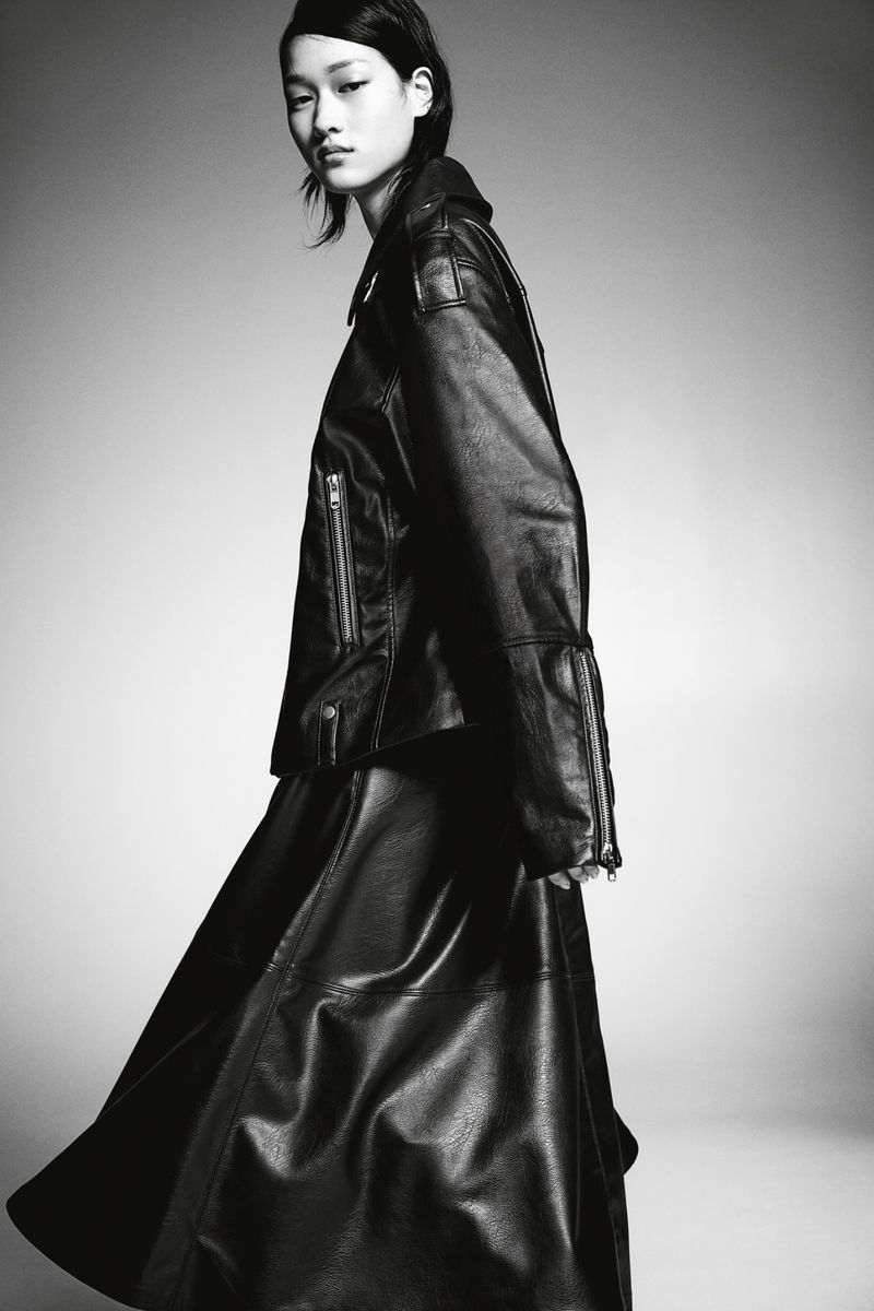 Monochrome portrait of an Asian model in a leather jacket and skirt against a gray background, showcasing fashion and style.