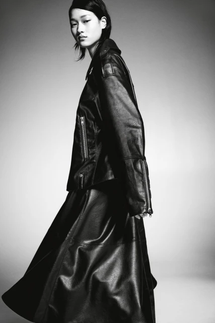 Monochrome portrait of an Asian model in a leather jacket and skirt against a gray background, showcasing fashion and style.