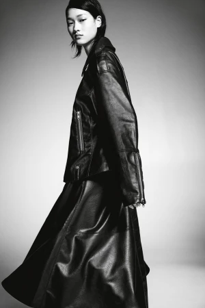 Monochrome portrait of an Asian model in a leather jacket and skirt against a gray background, showcasing fashion and style.