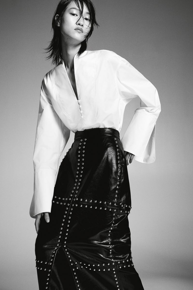 Monochrome portrait of a slender Asian fashion model wearing a white blouse and studded black leather skirt against a neutral background.