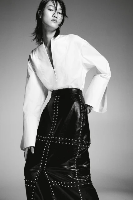 Monochrome portrait of a slender Asian fashion model wearing a white blouse and studded black leather skirt against a neutral background.