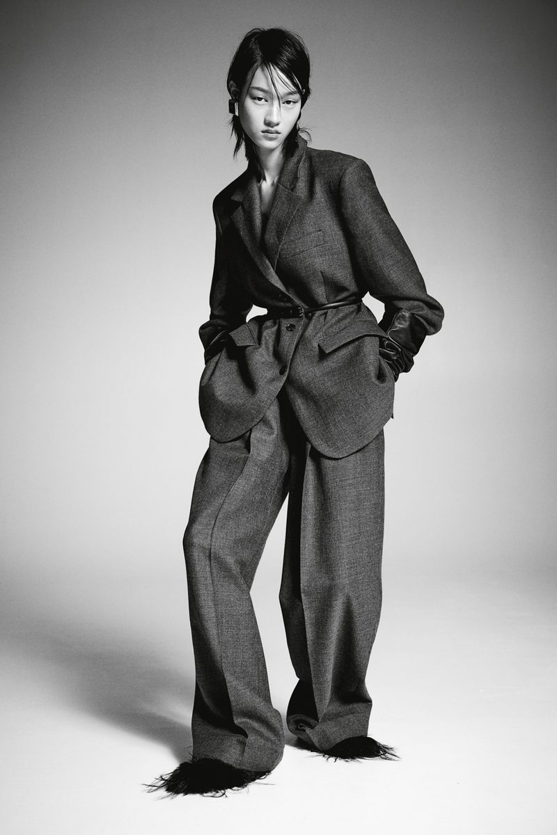 A full-length studio shot of an Asian woman in an oversized grey pantsuit with black trim at the hem of the pants, against a grey backdrop.