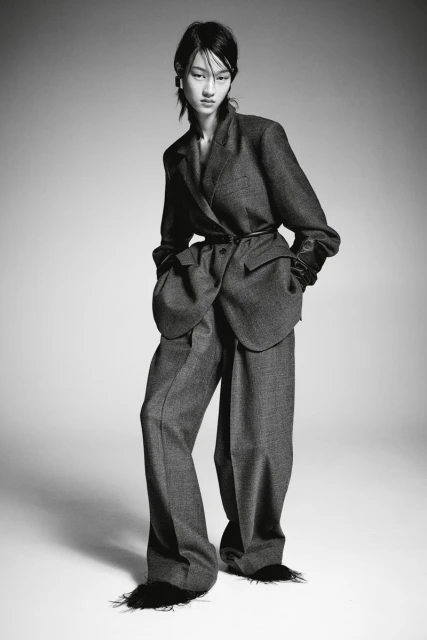 A full-length studio shot of an Asian woman in an oversized grey pantsuit with black trim at the hem of the pants, against a grey backdrop.