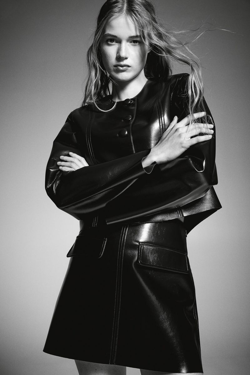 Black and white fashion portrait of a young adult female wearing a black, shiny, structured leather-look jacket and mini skirt.