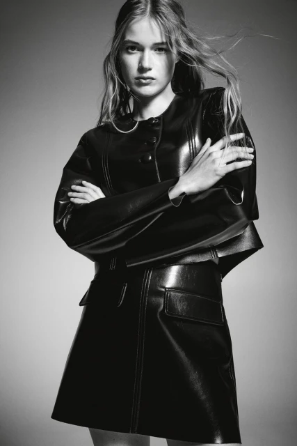 Black and white fashion portrait of a young adult female wearing a black, shiny, structured leather-look jacket and mini skirt.