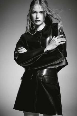 Black and white fashion portrait of a young adult female wearing a black, shiny, structured leather-look jacket and mini skirt.