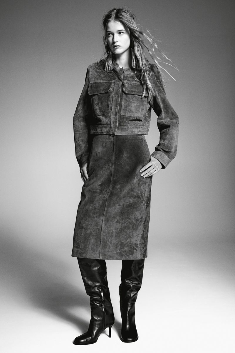 Monochrome fashion shot of a woman in a suede jacket, skirt, and boots, standing in a studio with soft lighting, neutral background.