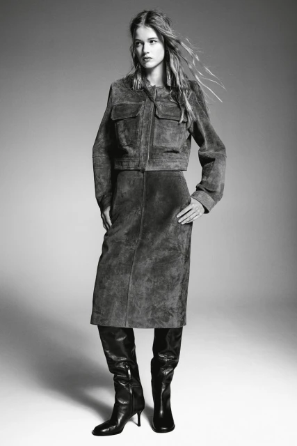 Monochrome fashion shot of a woman in a suede jacket, skirt, and boots, standing in a studio with soft lighting, neutral background.