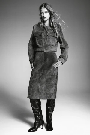 Monochrome fashion shot of a woman in a suede jacket, skirt, and boots, standing in a studio with soft lighting, neutral background.