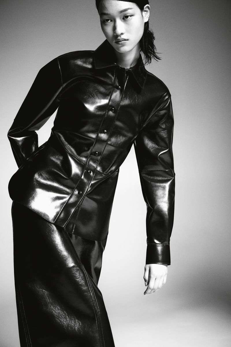 Monochrome portrait of a female model in a shiny black, long-sleeved shirt and matching skirt, set against a plain grey background.